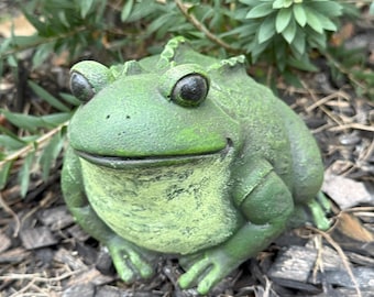 Könnte beinhalten: Eine grüne Froschstatue mit einem lächelnden Gesicht. Der Frosch hat große, dunkle Augen und sitzt auf einem Bett aus Mulch. Eine Gartendekoration.