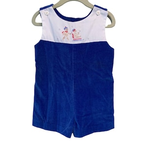 Puede incluir: Romper de pana azul con canesú blanco con una escena invernal bordada de dos figuras en un trineo. El romper tiene botones blancos en los hombros y se muestra en una percha.