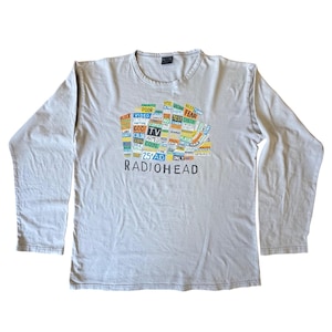 Può includere: Una maglietta a maniche lunghe grigio chiaro con un design grafico. Il design include la parola "RADIOHEAD" e un collage di parole e frasi colorate. La maglietta ha uno scollo tondo.