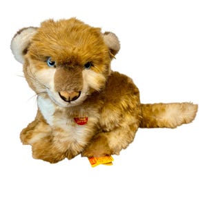 May include: A plush lion cub toy with brown and tan fur, blue eyes, and a white chest. The toy has a red tag with white text and a yellow tag with black text.