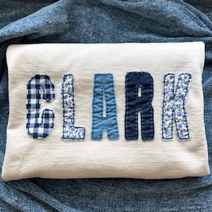 以下が含まれることがあります： 前面に「CLARK」の文字がアップリケされたクリーム色のスウェットシャツ。文字は、ギンガムや花柄など、さまざまなブルーの色合いと柄の生地でできています。スウェットシャツは、青色のテクスチャードブランケットの上に表示されています。