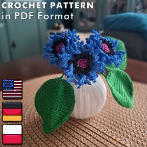 May include: Crochet pattern in PDF format. A crocheted bouquet of blue flowers with black and pink centers in a white vase. The flowers are surrounded by two green crocheted leaves.