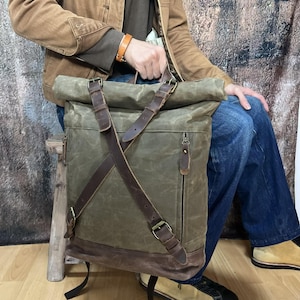 Pode incluir: Uma mochila de lona e couro marrom com fecho de rolo e alças ajustáveis. A mochila tem uma guarnição de couro marrom e um bolso com zíper na frente.