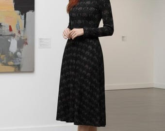 Puede incluir: Vestido negro de manga larga con estampado floral. El vestido tiene cuello redondo y falda acampanada. La persona lleva tacones negros. El fondo incluye una pintura y una pared blanca.
