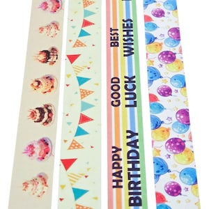 May include: Decorative washi tape rolls with various designs. One roll features birthday cakes, another has colorful pennants, and a third displays the text "Happy Birthday, Good Luck, Best Wishes." The last roll has balloons.