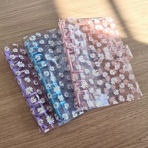 May include: Three clear plastic binders with daisy patterns. The binders are in shades of pink, purple, and blue. The binders have metal rings for holding papers.