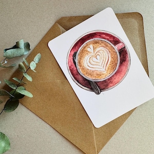 Könnte beinhalten: Aquarell-Illustration einer Kaffeetasse mit Latte Art in Herzform, auf einer roten Untertasse mit einem Löffel. Das Bild befindet sich auf einer weißen Karte, die auf einem braunen Umschlag liegt. Ein Eukalyptuszweig ist im Hintergrund.