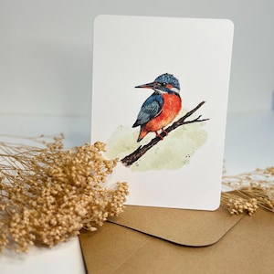 Könnte beinhalten: Aquarell-Grußkarte mit einem Eisvogel auf einem Ast. Der Vogel hat blaue, orangefarbene und weiße Federn. Die Karte befindet sich auf einem weißen Hintergrund, mit einem braunen Umschlag und getrockneten Blumen.