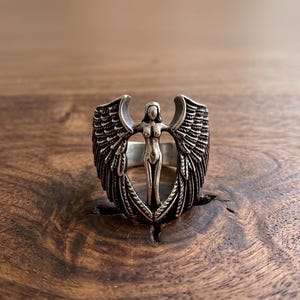 May include: A silver ring featuring a central figure with outstretched arms and detailed wings. The ring's design showcases an angel-like form, with intricate feather detailing on the wings. The ring is set against a dark wooden surface.