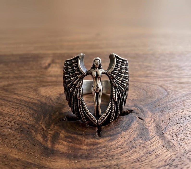 May include: A silver ring featuring a central figure with outstretched arms and detailed wings. The ring's design showcases an angel-like form, with intricate feather detailing on the wings. The ring is set against a dark wooden surface.