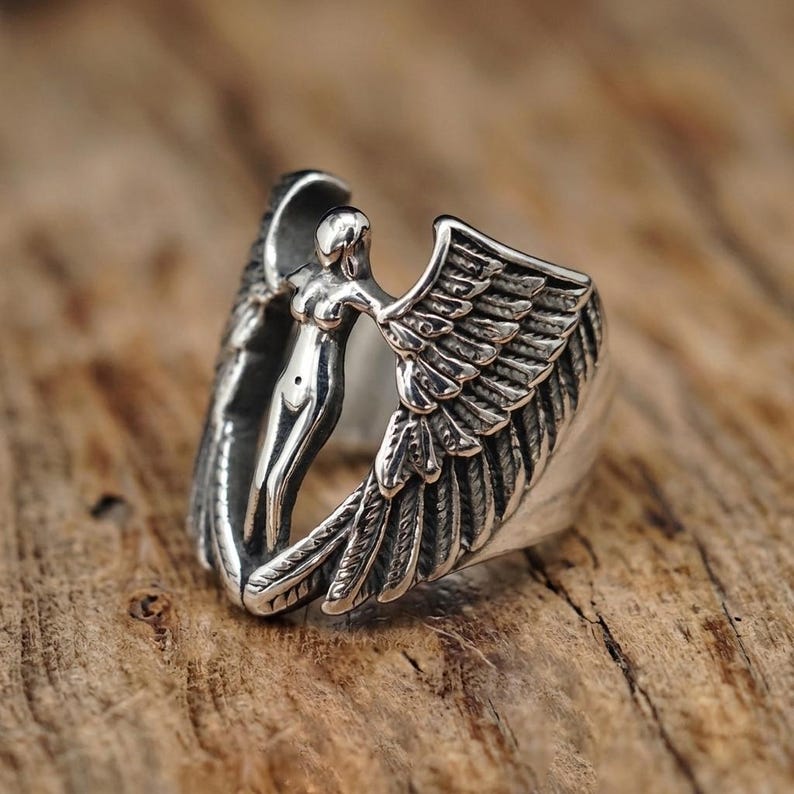 May include: A silver ring featuring a sculpted figure with outstretched wings. The ring's design includes detailed feathering on the wings and a smooth, polished finish on the figure, set against a wooden background.