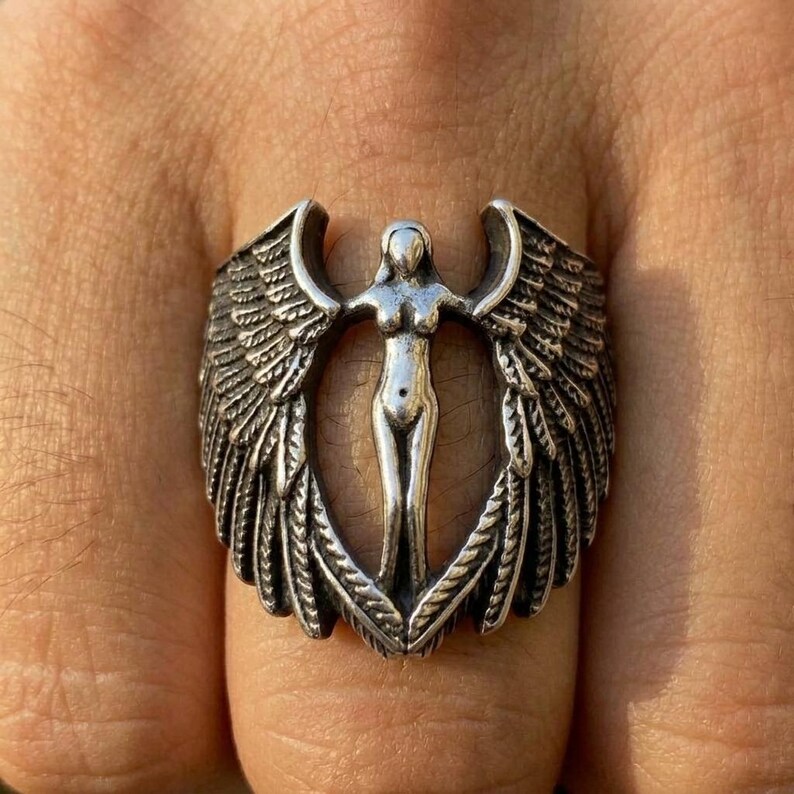 May include: Silver-toned ring featuring a central figure with outstretched arms, framed by detailed wings. The ring's design is intricate, with a focus on the feathered texture of the wings and the form of the central figure. The ring is worn on a finger.