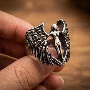 May include: Silver-toned ring featuring a sculpted angel figure with outstretched wings. The ring's design includes detailed feather textures on the wings and a stylized female form at the center. The ring is held against a blurred wooden background.