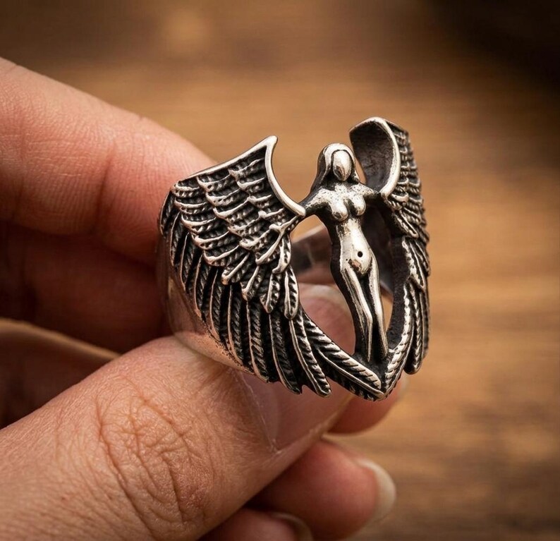 May include: Silver-toned ring featuring a sculpted angel figure with outstretched wings. The ring's design includes detailed feather textures on the wings and a stylized female form at the center. The ring is held against a blurred wooden background.