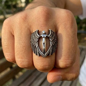 May include: A silver-colored ring featuring a central female figure with outstretched arms, framed by detailed, layered wings. The ring has an antique finish, showcasing intricate feather detailing and a bold, statement design.