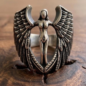 May include: A silver ring featuring a central figure of a woman with outstretched arms, flanked by detailed wings. The ring has a vintage, antique appearance, with intricate detailing on the wings and the woman's form. The ring is set against a wooden surface.