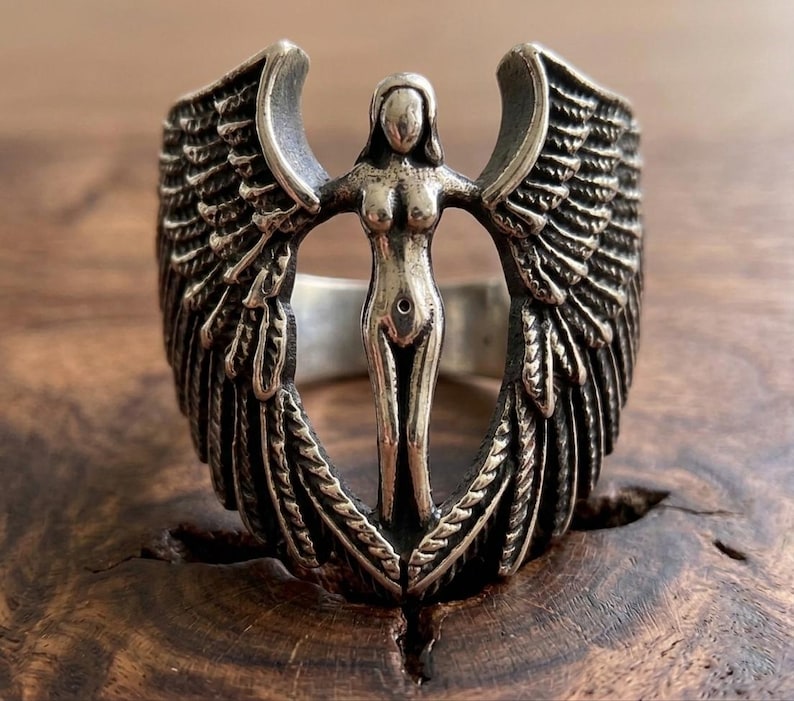May include: A silver ring featuring a central figure of a woman with outstretched arms, flanked by detailed wings. The ring has a vintage, antique appearance, with intricate detailing on the wings and the woman's form. The ring is set against a wooden surface.