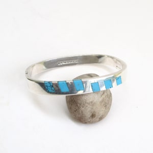 May include: A silver-tone hinged bangle bracelet with inlaid turquoise stones. The bracelet features a rectangular shape with a series of turquoise rectangles and a cluster of turquoise stones. The bracelet is displayed on a small, round, gray stone.