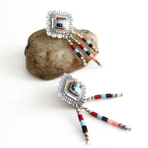 May include: A pair of silver earrings with a square, multi-colored stone center and dangling beaded strands. The earrings have a detailed, scalloped edge design. The beads are red, black, turquoise, and pink.