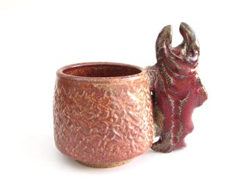 May include: A ceramic mug with a red and brown glaze. The mug has a unique handle shaped like a fish.