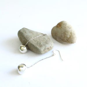 May include: A pair of silver-toned drop earrings featuring a delicate chain attached to a smooth, polished spherical bead. Each earring includes a curved hook for attachment, showcasing a reflective metallic finish and a simple, elongated design.