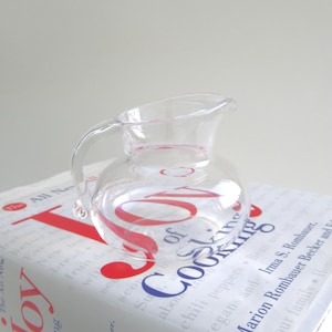 May include: A clear glass pitcher with a handle, sitting on top of a white book with red and blue text that reads "The Joy of Cooking".