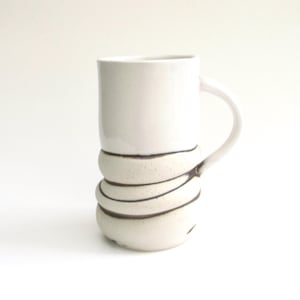 May include: A white ceramic mug with a cylindrical upper section and a lower section wrapped in a textured, stacked design with dark brown lines. The mug has a curved handle and sits on a white surface.