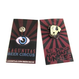 May include: A white and brown dog head enamel pin with a black collar and multiple black eyes. The pin is on a red and black background with the text "See things you can't un-see". The pin is packaged on a card with the Lagunitas Beer Circus logo and website.