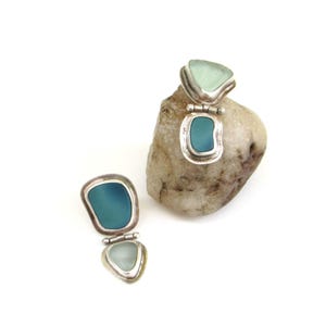 May include: Two pairs of handcrafted earrings with a silver setting and sea glass in shades of teal and light blue. One pair is open, revealing the sea glass, while the other is closed. The earrings are displayed on a small, light-colored rock.