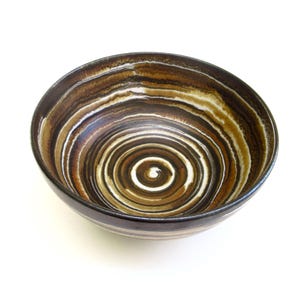 May include: A ceramic bowl with a spiral design in shades of brown, tan, and white. The bowl's interior features concentric circles, creating a visually appealing pattern. The rim and exterior base are a darker brown.
