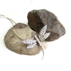 May include: A silver-toned dragonfly pendant necklace featuring clear, sparkling stones embedded across the wings and body. The pendant is attached to a thin, delicate silver-toned chain that rests against the textured surface of two natural stones.