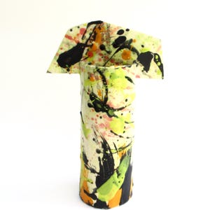 May include: A cylindrical ceramic vase with an irregular, flared top edge. The surface is off-white and features a vibrant, multi-colored paint splatter pattern in shades of black, bright lime green, orange, and soft pink, creating a bold visual effect.
