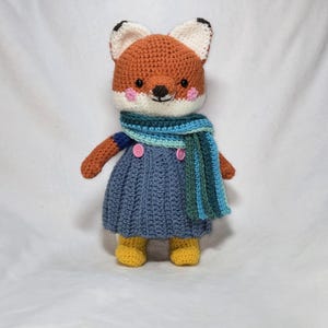 May include: A crocheted fox plush toy wearing a blue dress with buttons and a striped scarf. The fox has orange fur, white accents, and pink cheeks.