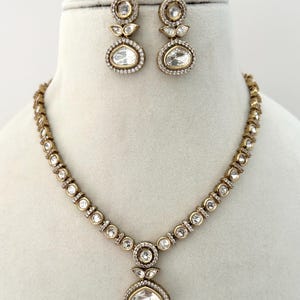 Peut inclure: Un ensemble collier et boucles d'oreilles de couleur dorée. Le collier présente une série de pierres rondes et transparentes reliées par des maillons dorés, avec un plus grand pendentif au centre. Les boucles d'oreilles reprennent le design du pendentif.