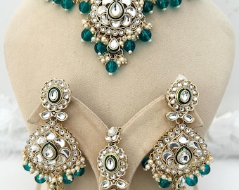 May include: An ornate gold-toned jewelry set featuring a choker necklace, earrings, and a pendant. The set includes clear and emerald green gemstones, and small white pearls. The necklace and earrings have a floral design.