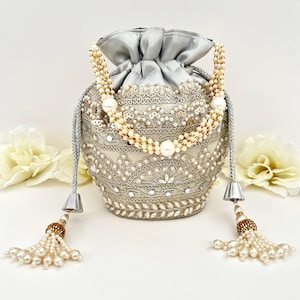 May include: A silver drawstring bag with intricate embroidery and embellishments. The bag features a pearl-beaded handle and tassel, with a silver satin top. The bag is decorated with small mirrors and pearls.