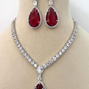 May include: A jewellery set comprising a necklace and matching earrings. The necklace and earrings have a silver-coloured metal setting. The earrings and necklace pendant feature a large, teardrop-shaped, deep red stone surrounded by smaller, clear stones.