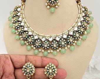 Puede incluir: Un conjunto de joyas ornamentado que incluye un collar, pendientes y una pieza para la cabeza. El conjunto tiene un acabado dorado con detalles verdes y blancos, incluyendo pequeñas cuentas y diseños en forma de lágrima. Los pendientes son en forma de campana.