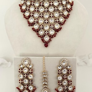 Op de afbeelding: Een sierlijke, goudkleurige sieradenset met een choker-ketting, oorbellen en een hoofdband. De set is versierd met heldere, facetgeslepen stenen en kleine, ronde rode kralen. De ketting en oorbellen hebben een druppelvormig ontwerp.