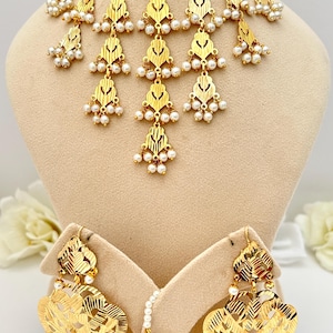May include: A gold-colored jewelry set featuring a necklace and matching earrings. The necklace and earrings have a floral design with dangling elements and small white pearls. The jewelry is displayed on a mannequin.