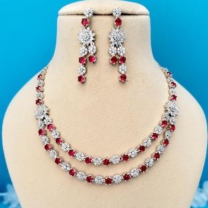 May include: A silver-toned necklace and earring set with clear and red gemstones. The necklace is a two-strand design. The earrings are a dangling style with a floral pattern. The jewellery is displayed on a mannequin against a blue background.