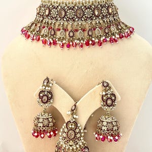 May include: An ornate, gold-toned jewelry set featuring a choker necklace, earrings, and a headpiece. The set is adorned with burgundy-colored stones and small white beads, with dangling accents. The jewelry is displayed on a mannequin.