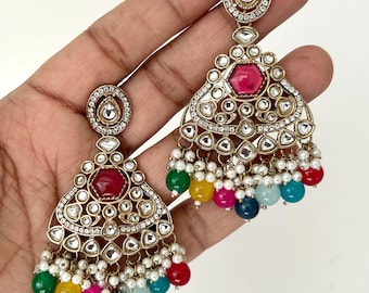May include: Gold-toned chandelier earrings with a geometric design. Each earring features a central red gemstone, surrounded by clear crystals and white pearls. Colorful beads in green, yellow, pink, blue, and red dangle from the bottom.