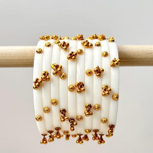 May include: A stack of white bangles with vertical grooves and gold-coloured embellishments. The gold accents include small, clustered beads and individual round beads, creating a textured and ornate appearance. The bangles are displayed on a wooden dowel.
