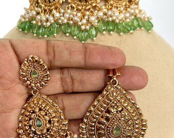 May include: Gold-toned jewelry set featuring a necklace and matching earrings. The necklace and earrings have intricate detailing, green gemstone accents, and white pearl and green bead dangles. The set is displayed on a mannequin head.