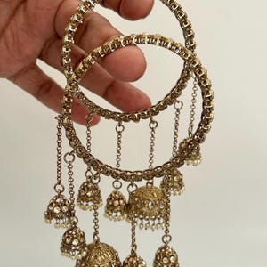 May include: Two gold bangles with detailed patterns and hanging bell charms. Each bangle has a chain-linked design and small bells with faux pearl details. The jewellery is held against a plain background.