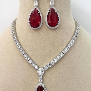 Peut inclure: Un ensemble de bijoux éblouissant comprenant un collier et des boucles d'oreilles assorties. Le collier et les boucles d'oreilles ont un sertissage en métal argenté, orné de pierres claires et étincelantes. Les boucles d'oreilles et le pendentif du collier présentent de grandes pierres précieuses rouges en forme de goutte.