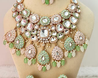 May include: A three-piece jewelry set featuring a necklace, earrings, and a headpiece. The set has a gold-tone metal base with pink and mint green accents, clear stones, and dangling green and pink beads. The necklace and earrings are displayed on a mannequin.