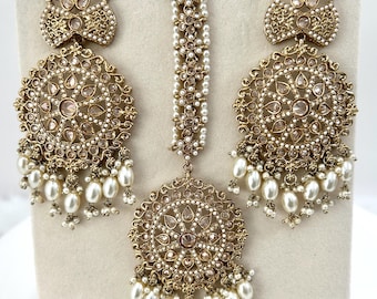 May include: A gold-toned jewelry set featuring a pair of large, round earrings and a headpiece. The earrings and headpiece are adorned with small, clear stones and dangling white pearls. The set is displayed on a neutral-colored background.