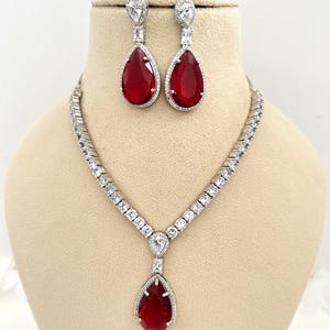 May include: A silver-coloured jewellery set comprising a necklace and matching earrings. The necklace and earrings feature a teardrop-shaped, deep red gemstone surrounded by clear crystals. The earrings are drop style, and the necklace has a similar pendant.